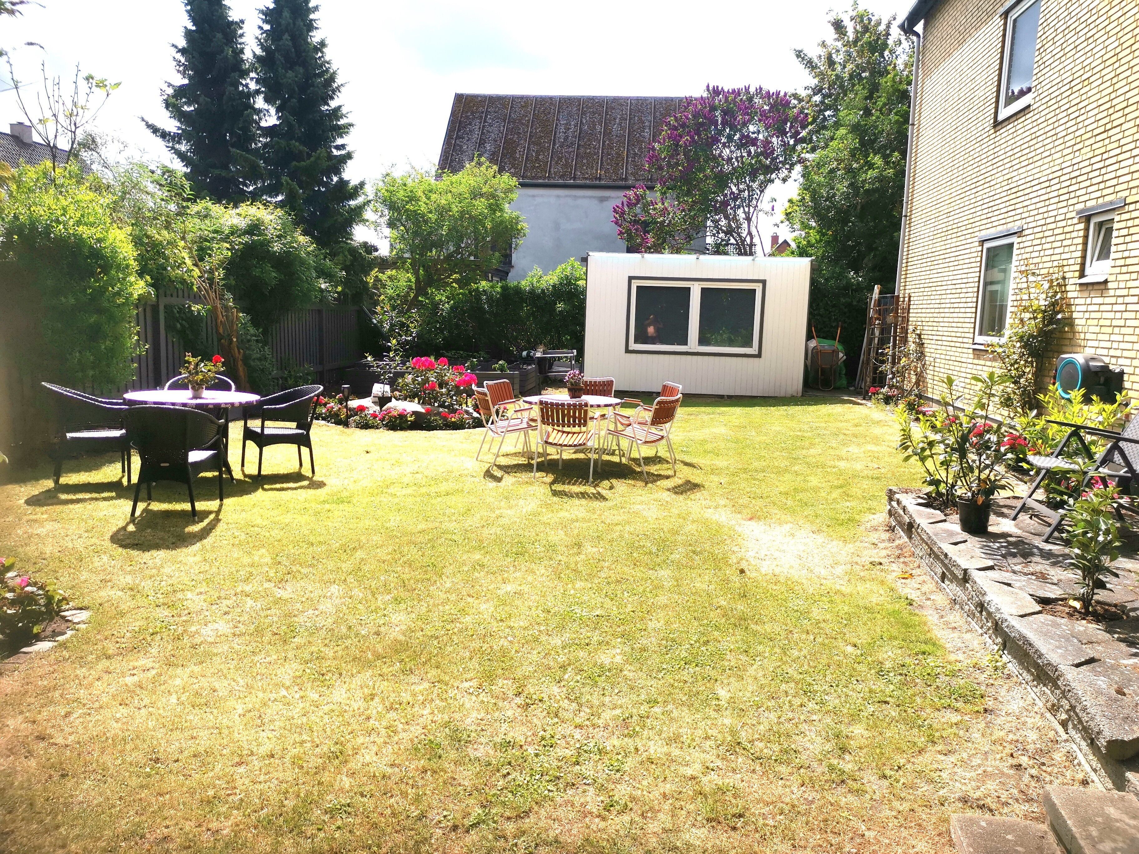 Ein Garten mit Terrasse, einem kleinen weißen Gebäude und verschiedenen Pflanzen.