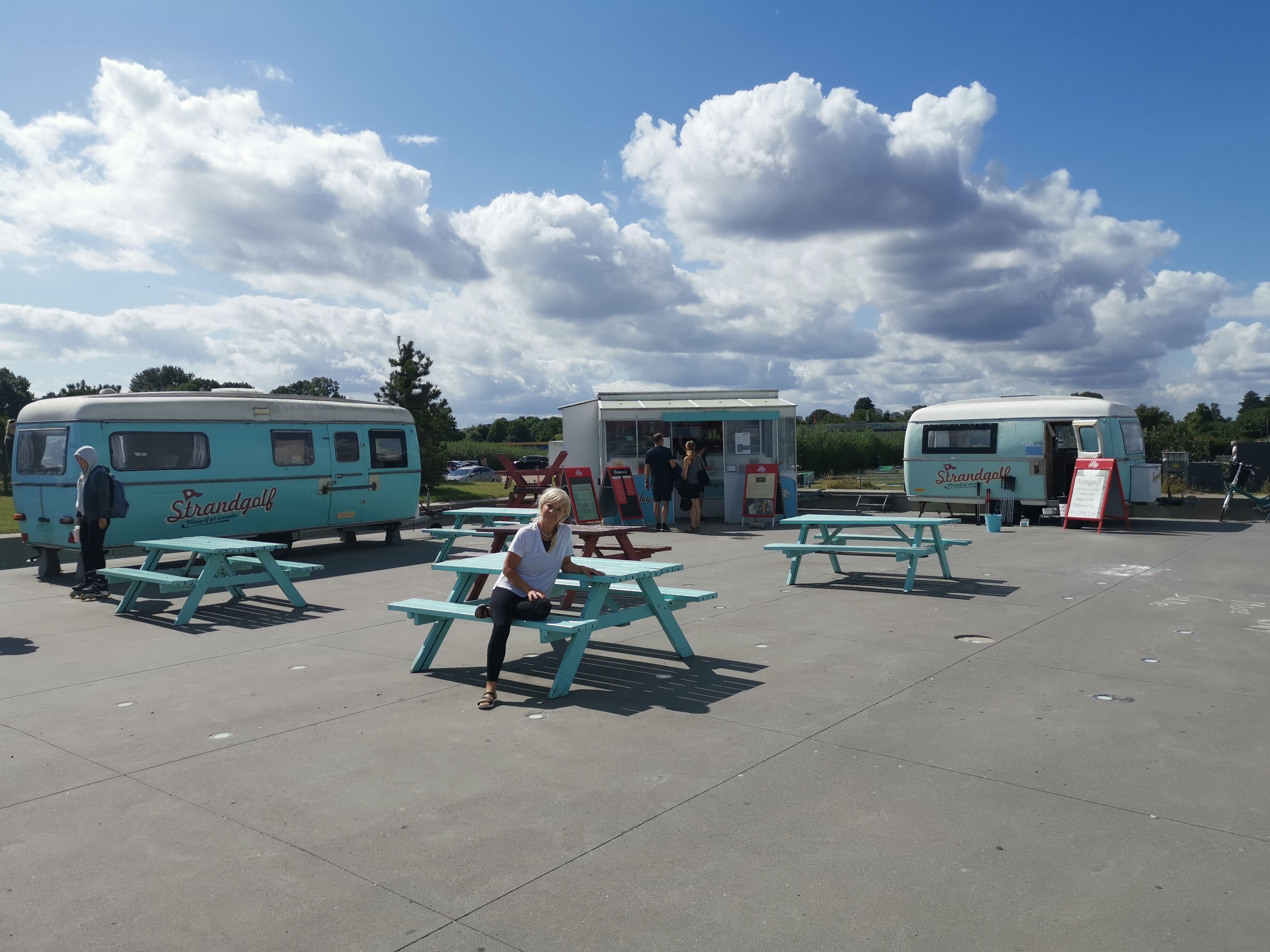 A parking area with vintage-style mobile food service units, picnic tables, and a clear sky.