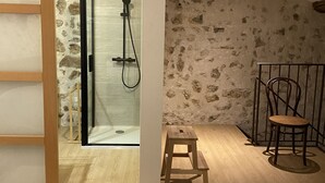 Ein Schlafzimmer mit einem Bett, einem hölzernen Hocker, einem Stuhl, einem kleinen Tisch und einem Duschbereich mit Glastür.