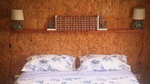 Een bed met een blauw-witte bloemenlaken, twee kussens, een houten plank erboven en twee wandlampen aan weerszijden.