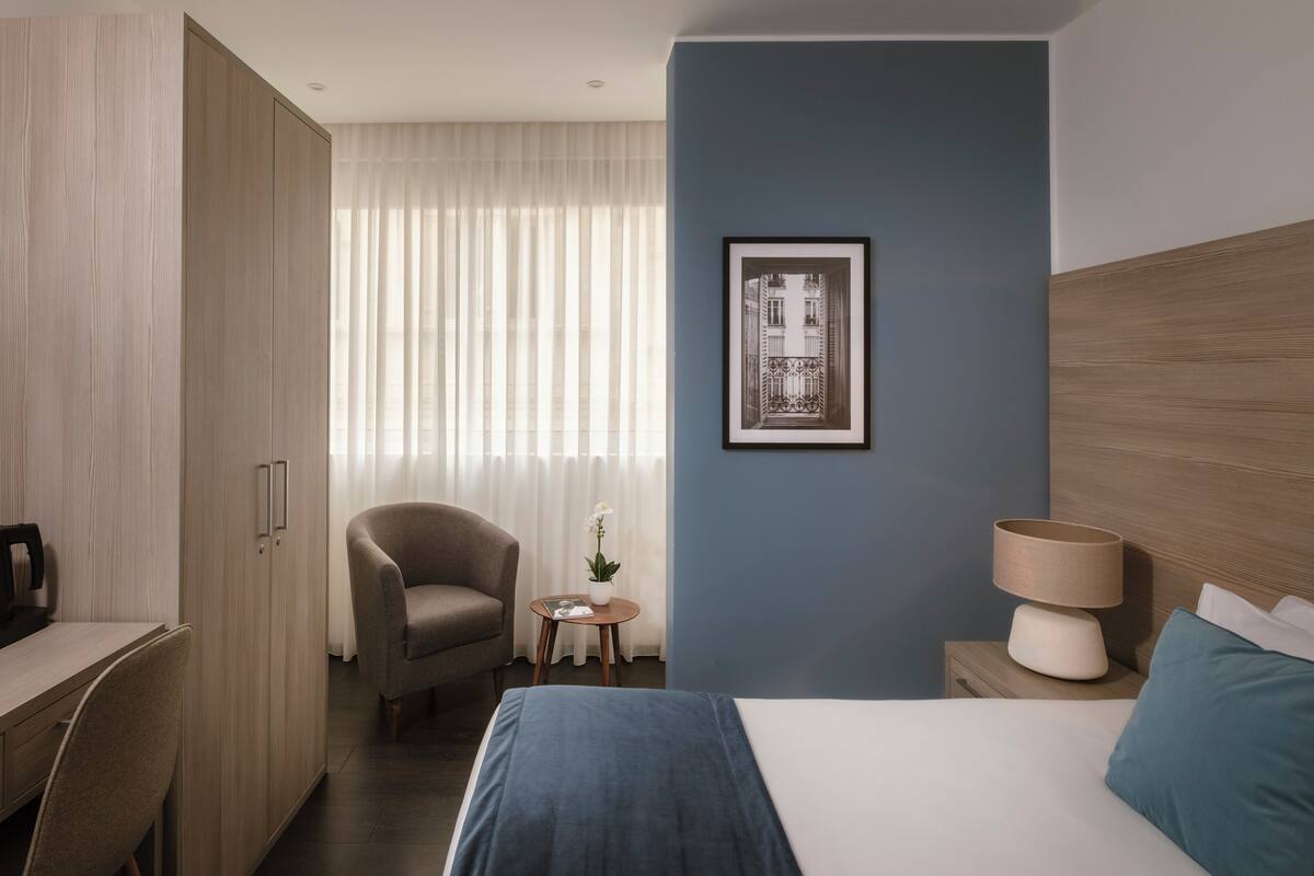 A hotel room with a bed, a desk, a chair, a nightstand, a lamp, a framed picture on the wall, and a window with curtains.