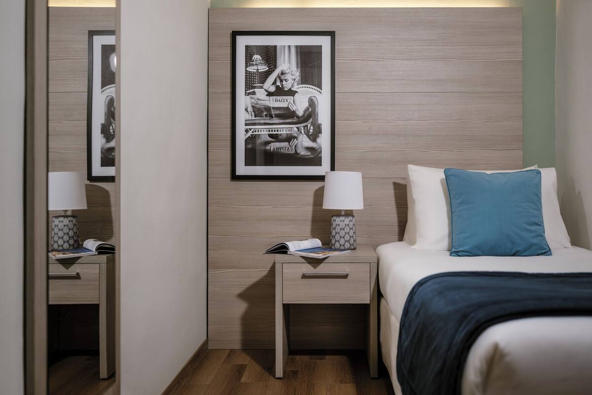 A hotel room with a bed, a nightstand with a lamp, a framed picture on the wall, and a wooden panel design.