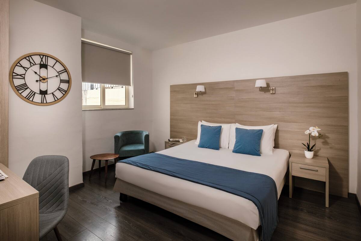 A modern hotel room with a large bed, a desk, a chair, a clock, and a window.