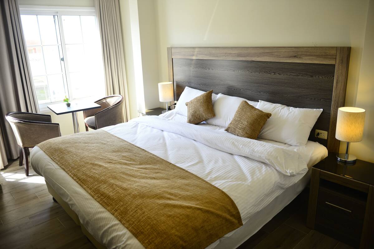 A hotel room with a large bed, a small table, a chair, and a nightstand with a lamp.