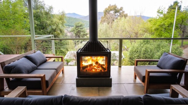 A wood-burning stove with a fire, surrounded by wooden furniture and a balcony with a view of trees and hills.