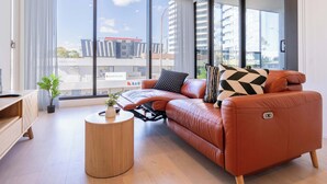 A modern living room with a leather sofa, a round wooden coffee table, and a large window offering a city view. - Modern Luxury 2B2B Sky Garden SmartTV (Glen Waverley)
