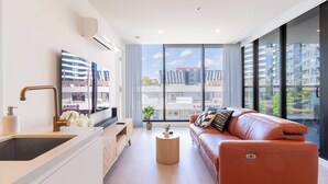 A modern living room with a sofa, a coffee table, a TV, and a large window with a city view. - Modern Luxury 2B2B Sky Garden SmartTV (Glen Waverley)