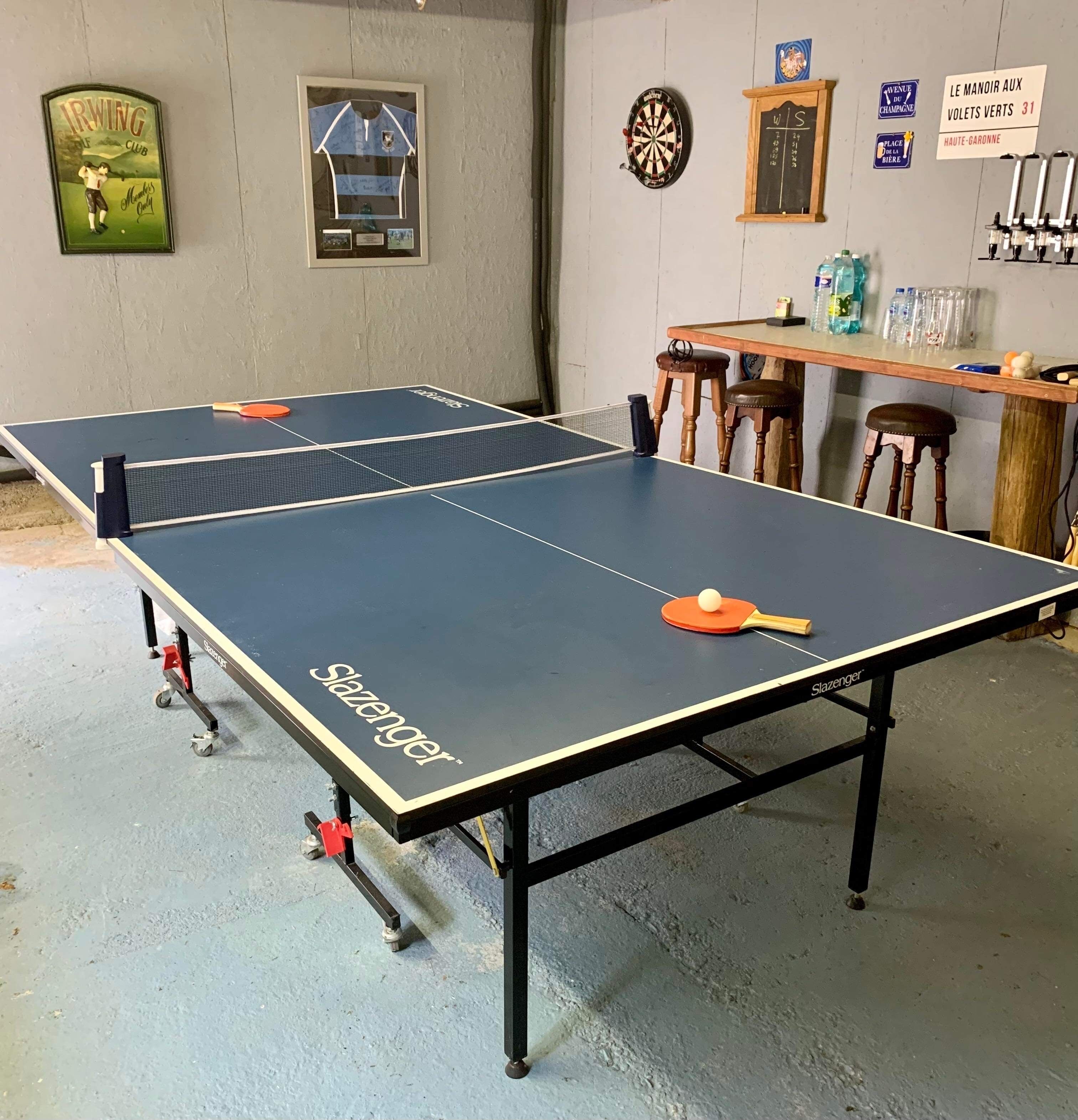 A ping pong table with a Slazenger logo, a dartboard, and a framed picture of a golf club on the wall.