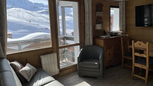 Een knus interieur met houtaccenten, een bank, een stoel, een eettafel en een uitzicht op besneeuwde bergen door grote ramen.