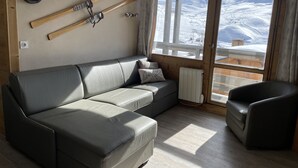 Een moderne woonkamer met een grijze leren bank, een houten stoel en een groot raam met uitzicht op een besneeuwd landschap.