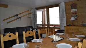Een zithoek met een houten tafel gedekt voor vier personen, een zwarte bank, houten stoelen en een houten wand met ski's en een bord met daarop een welkomstboodschap.