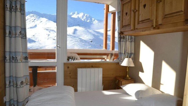 Een slaapkamer met een bed, nachtkastje, lamp en uitzicht op besneeuwde bergen door een raam.