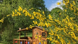 A wooden playhouse with a balcony, surrounded by lush greenery and yellow flowers.