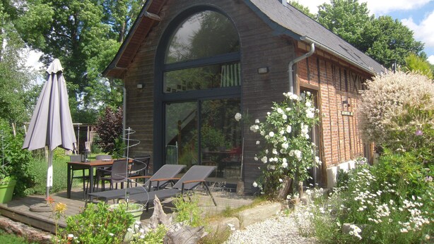 A wooden house with a large arched window, a deck with outdoor furniture, and a garden with flowers.