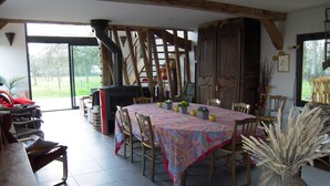 A dining area with a large table covered in a colorful tablecloth, surrounded by wooden chairs. There is a fireplace with a chimney, and a large wooden cabinet is visible in the background.