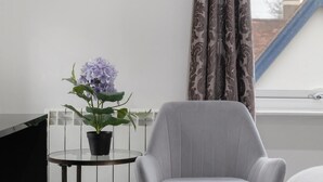 A room with a grey armchair, a small round table with a potted plant, and a window with patterned curtains.