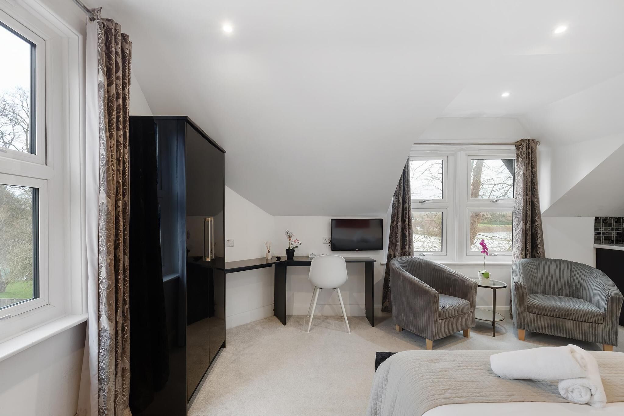 A modern hotel room with a large bed, two armchairs, a TV, and a desk with a small table.
