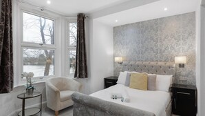 Een slaapkamer met een groot bed, twee nachtkastjes, een stoel, een kleine tafel met een vaas en een raam met gordijnen.