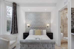 Een moderne slaapkamer met een gestoffeerd hoofdeinde, een groot bed met wit en grijze beddengoed, twee nachtkastjes met lampen, een raam met patroon gordijnen, en een badkamer met een douchecabine van glas.