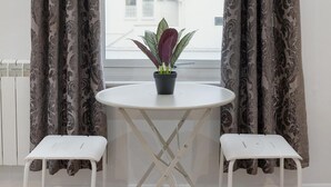 A room with a window, two white stools, a round table with a potted plant, and patterned curtains. - Central and Modern One Bedroom Flat (Oxford)