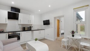 A modern kitchen with white cabinets, a built-in oven, and a flat-screen TV. There is a small dining area with a white table and bench. - Central and Modern One Bedroom Flat (Oxford)