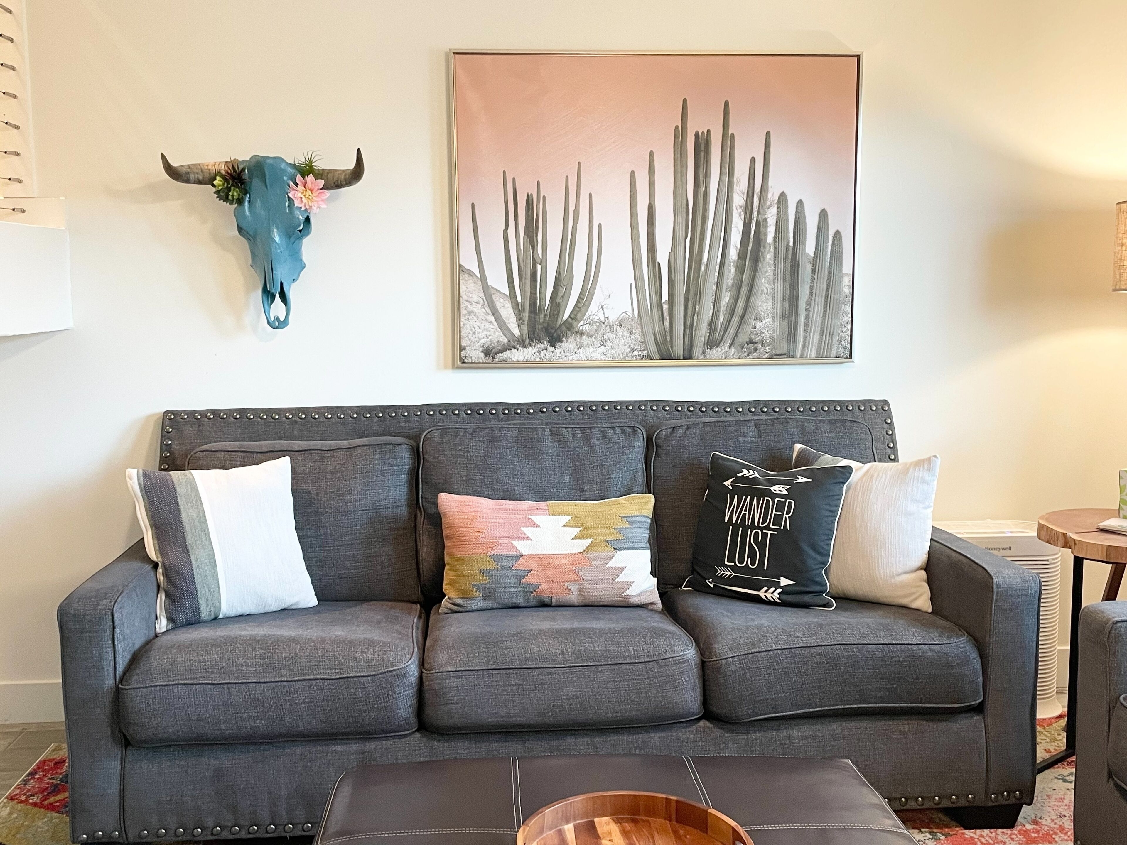A grey sofa with three cushions, a wall-mounted bull skull, and a cactus painting.
