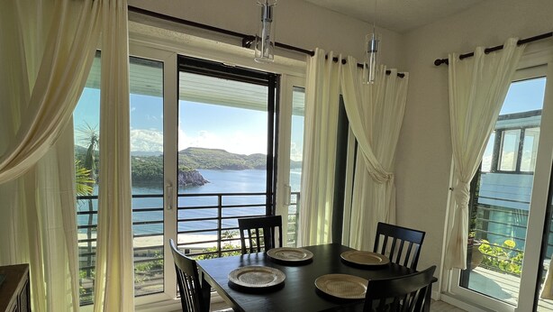 A dining area with a table and chairs, large windows with curtains, and a view of the sea. - Seaside Blue <br>Hosted by Shilpi Singh (St Thomas)