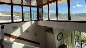 A room with large windows offering a view of a hilly landscape and a distant building. - Seaside Blue <br>Hosted by Shilpi Singh (St Thomas)