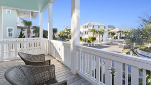 A wooden deck with wicker chairs, a white railing, and a view of a coastal town with buildings and palm trees. - Aqua Easy - Destin Pointe, Gulf Views, Lakefront, Free Beach Service, Cmty Pool! (Destin)