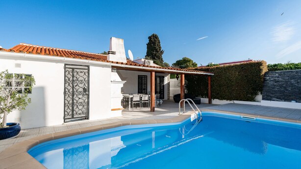 Una piscina con zona de patio, espacio de estar techado y un edificio blanco con tejado de tejas.