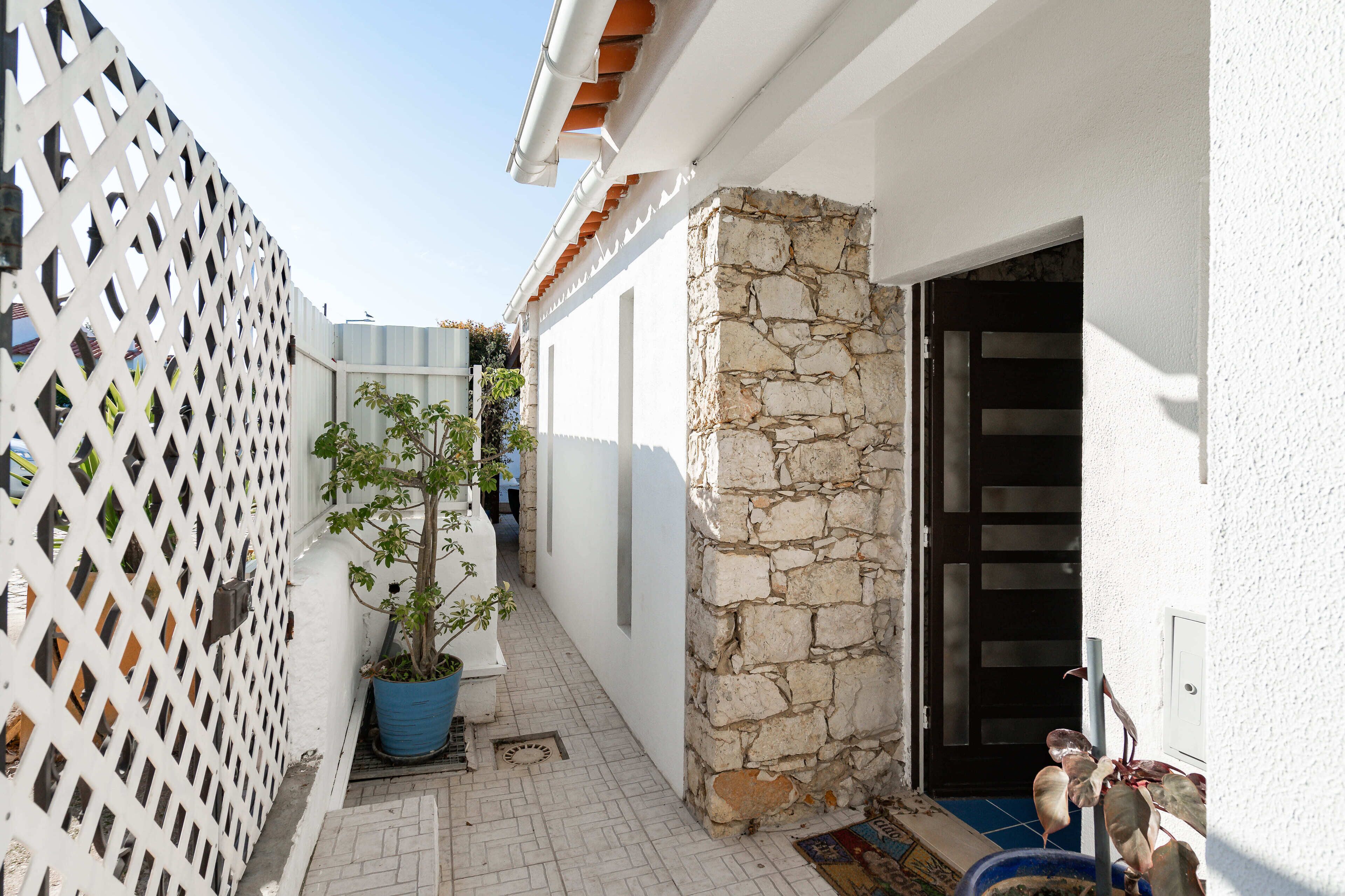 Un étroit couloir extérieur, bordé d’une clôture en treillis blanc, avec un pot de fleurs et un mur en pierre.