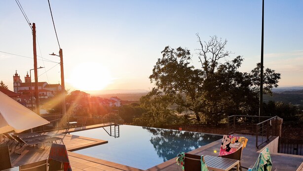 An infinity pool with a view of a sunset and distant buildings. - Casa Do Vidoeiro (Seia)