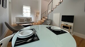A modern living room with a glass dining table set for two, a sofa, a staircase, and a flat-screen TV. - Casa Do Vidoeiro (Seia)