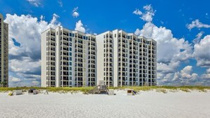 A beachfront high-rise hotel with multiple balconies, a sandy beach in the foreground, and a clear blue sky with clouds. - Salt and Sunlight: Spacious, airy 9th Floor Navarre Towers condo located on the Gulf! (Navarre)