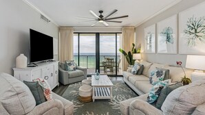 A spacious living room with a flat-screen TV, a ceiling fan, and a balcony with a view of the ocean. - Salt and Sunlight: Spacious, airy 9th Floor Navarre Towers condo located on the Gulf! (Navarre)