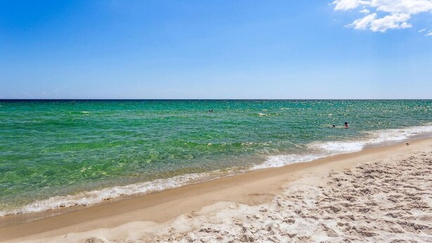 A beach with clear turquoise water, a sandy shore, and a few people swimming. - Salt and Sunlight: Spacious, airy 9th Floor Navarre Towers condo located on the Gulf! (Navarre)