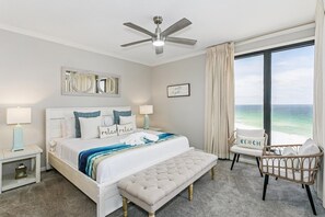 Una camera da letto con un letto, comodini, una panca, una sedia, una ventola a soffitto e un'ampia finestra vista oceano.
