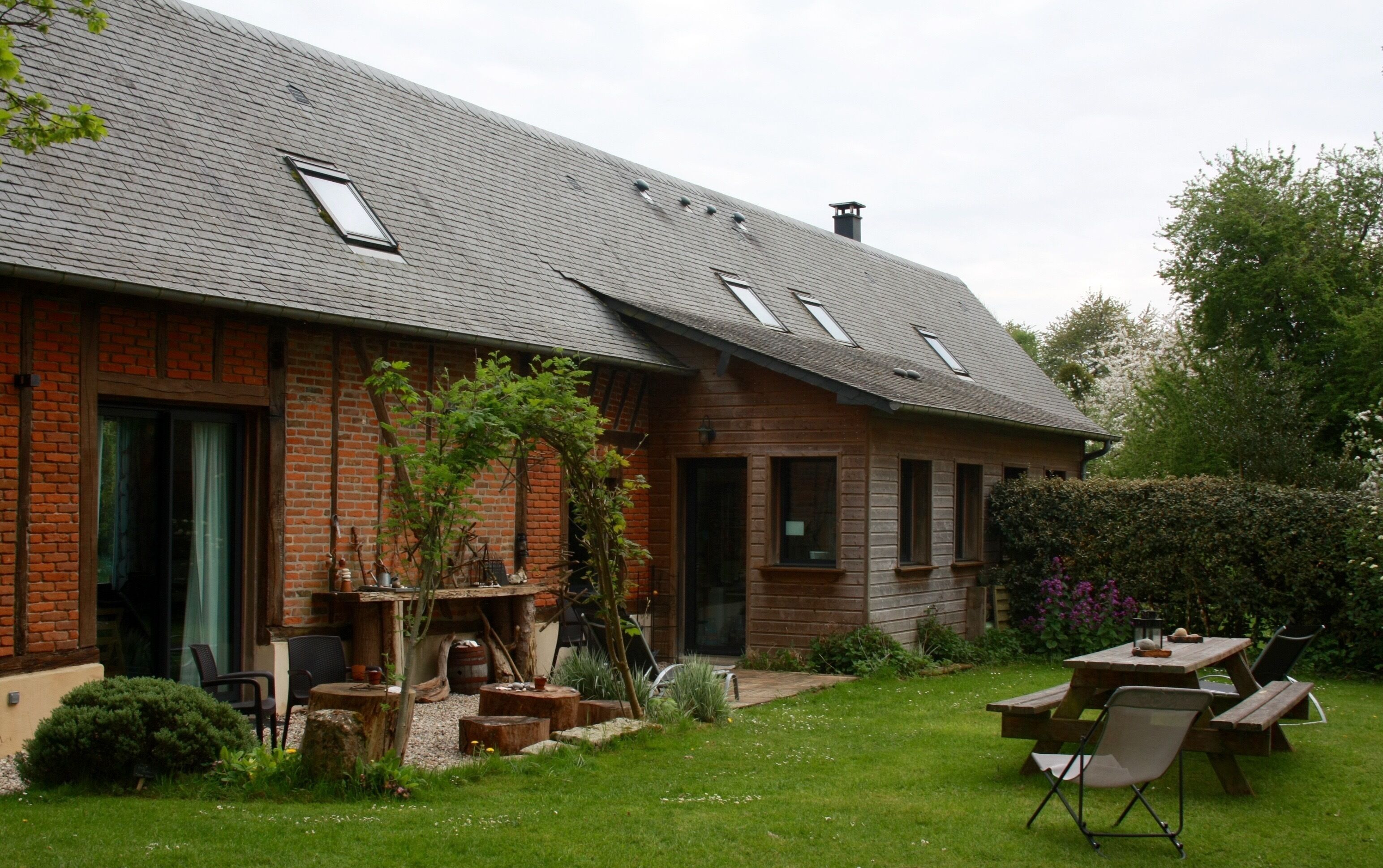 A brick house with a sloped roof, a patio with a table and chairs, and a garden with various plants and a small tree.