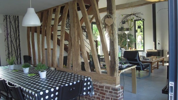 Un coin repas avec une table noire recouverte d’une nappe blanche à pois, entourée de poutres en bois et de plantes en pot.