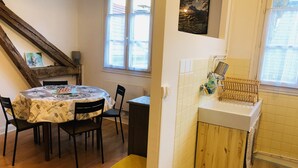 A dining area with a round table covered in a patterned tablecloth, wooden chairs, and a window with blinds. - Cosy, renovated family home in the heart of the Pyrenees (Eaux-Bonnes)