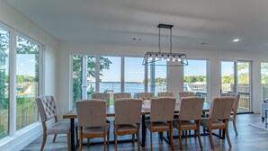 A spacious dining area with a large table and multiple chairs, large windows with a view of a body of water, and a modern pendant light fixture.