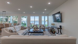 A spacious living room with a large sofa, a coffee table, and a TV mounted on the wall. There are multiple windows providing a view of the outdoors.