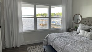 A bedroom with a large window overlooking a dock and water. There is a bed with white bedding, a bench, and a small table with a lamp.