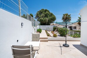 A patio with a white chair, a small table, and a metal umbrella. There is a white wall with a window and a door.