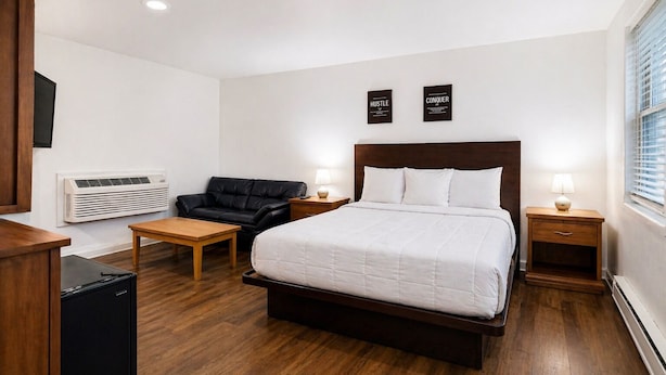 A hotel room with a large bed, a black leather sofa, a wooden table, two bedside tables with lamps, and a wall-mounted air conditioner. - Relax Inn Smyrna Dover Coastal Gateway (Smyrna)