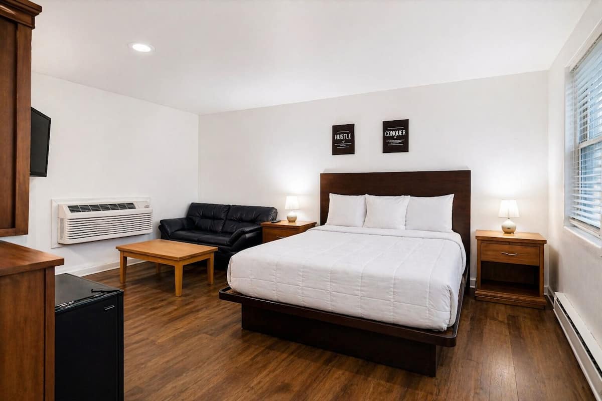 A hotel room with a large bed, a black leather sofa, a wooden table, two bedside tables with lamps, and a wall-mounted air conditioner.