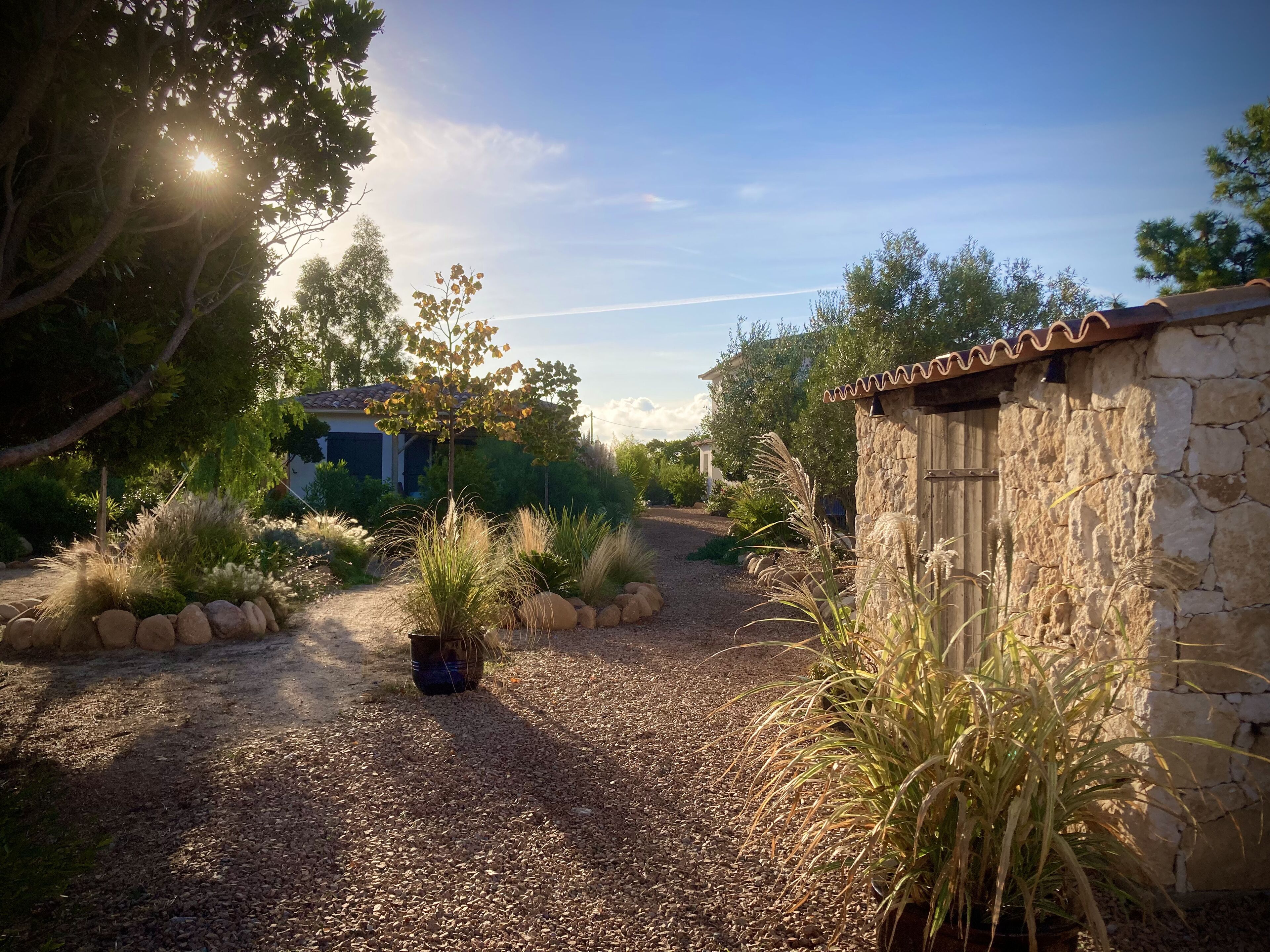 A gravel path leading to a stone building with a tiled roof, surrounded by various plants and trees.
