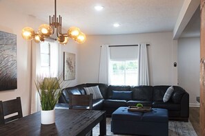 A modern living room with a dark leather sofa, a wooden dining table, and a chandelier.