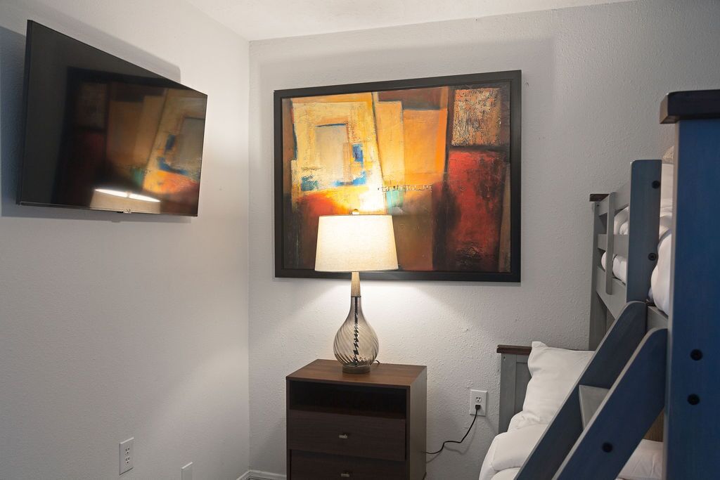 Ein Schlafzimmer mit einem Bett, einem Nachttisch mit Lampe, einem an der Wand montierten Fernseher und einem Gemälde über dem Bett.