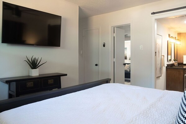 A hotel room with a bed, a flat-screen TV mounted on the wall, a small potted plant, a dresser with drawers, and a door leading to another room.